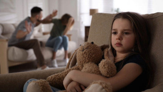 A young child clutches a teddy bear, listening to her parents argue in the next room